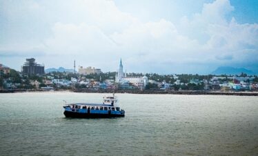 A passenger ship sails past the scenic waterfront of Kanyakumari, India, with urban landscapes in the background.