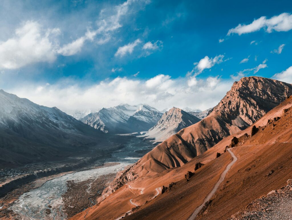Breathtaking aerial view of Spiti Valley's snow-capped mountains and winding roads under a vibrant blue sky.