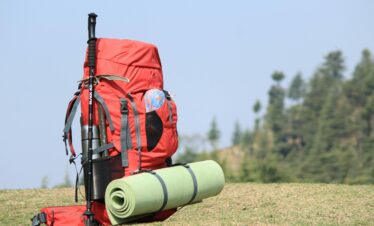 Red backpack with hiking pole and mat on grassy field with forest background.