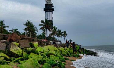 A stunning view of a lighthouse on the Indian coast, surrounded by palm trees and mossy rocks.