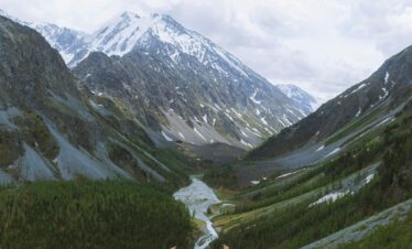 Hiker celebrating atop a mountain with breathtaking views of a lush valley and snow-capped peaks.
