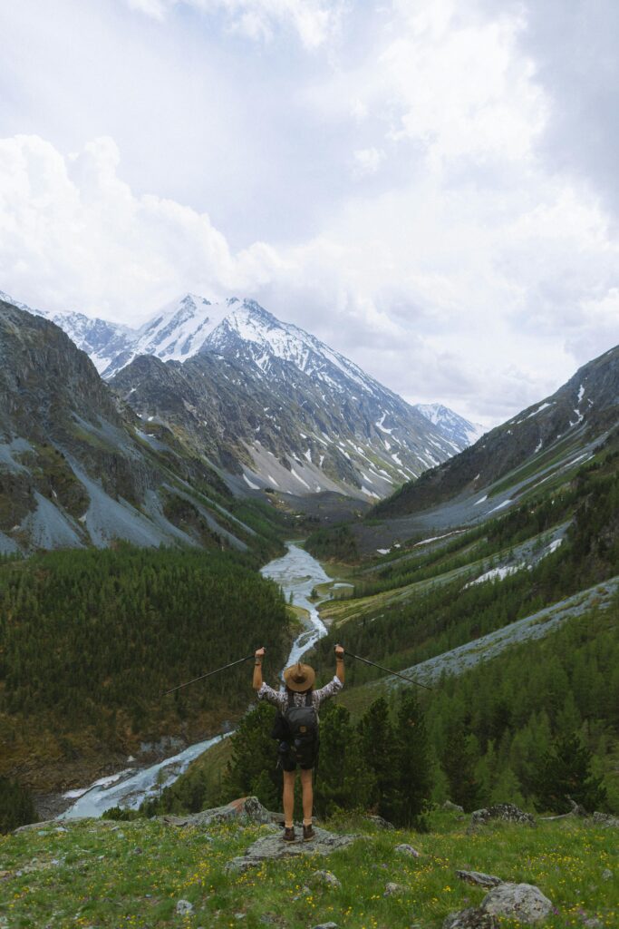 Hiker celebrating atop a mountain with breathtaking views of a lush valley and snow-capped peaks.
