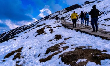 Group of hikers navigating snowy trails at Tungnath, India under a vibrant sky.