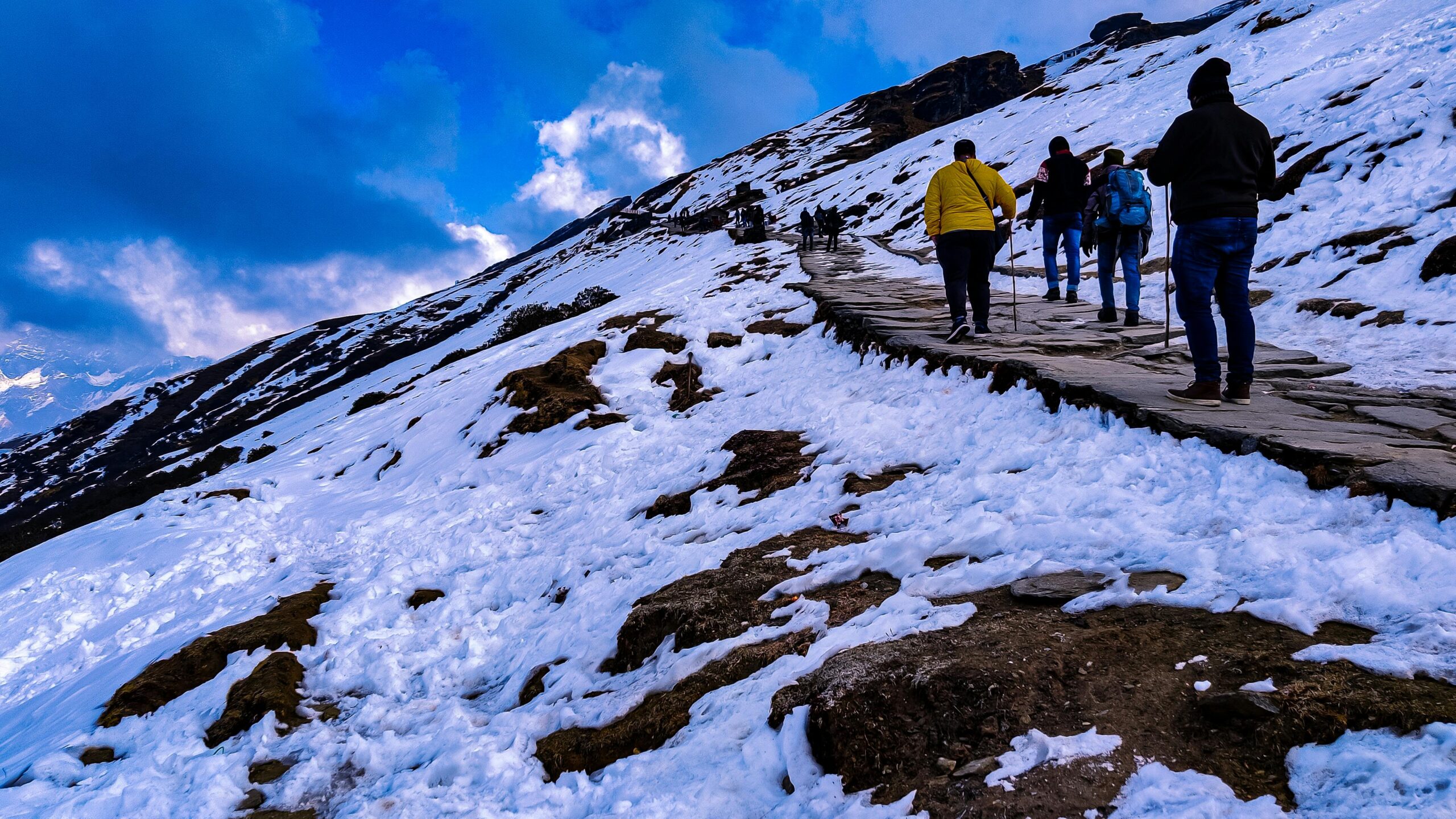 Group of hikers navigating snowy trails at Tungnath, India under a vibrant sky.